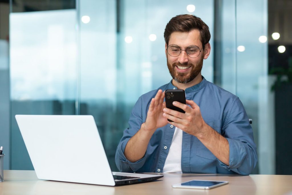 Homem no celular e com o computador ao lado