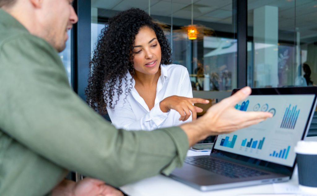 Business man and business woman in a meeting at the office. There is a laptop on the table showing finance charts and graphs. They are looking and pointing at the computer